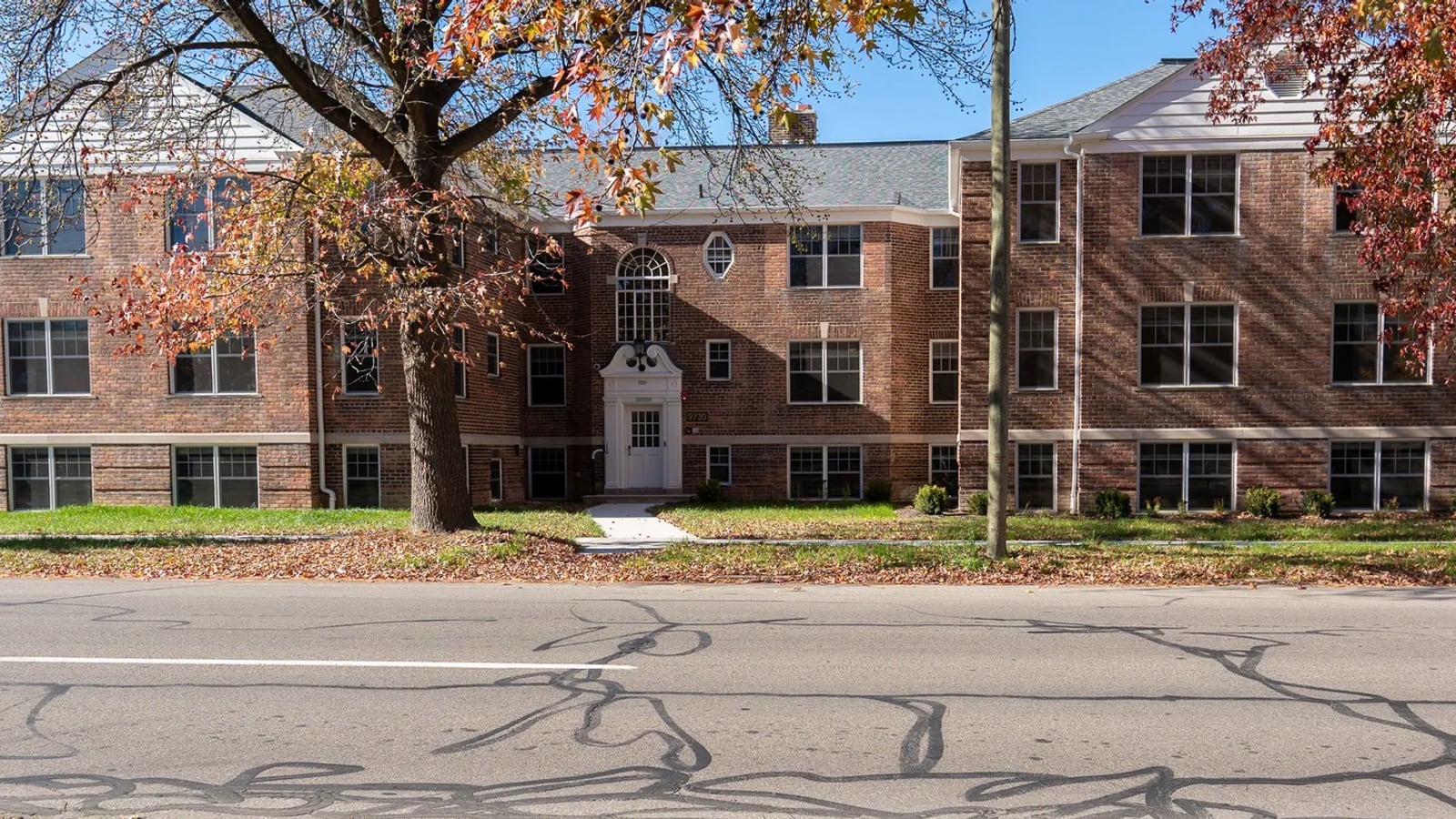 Photograph across a street of a long three-story red brick apartment building with white trim and a centered doorway, behind a lawn and autumn trees