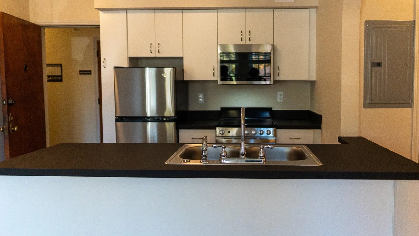 Photograph of an apartment kitchen with white cabinets, a dark counter with a double sink, a steel refrigerator, a range and a microwave