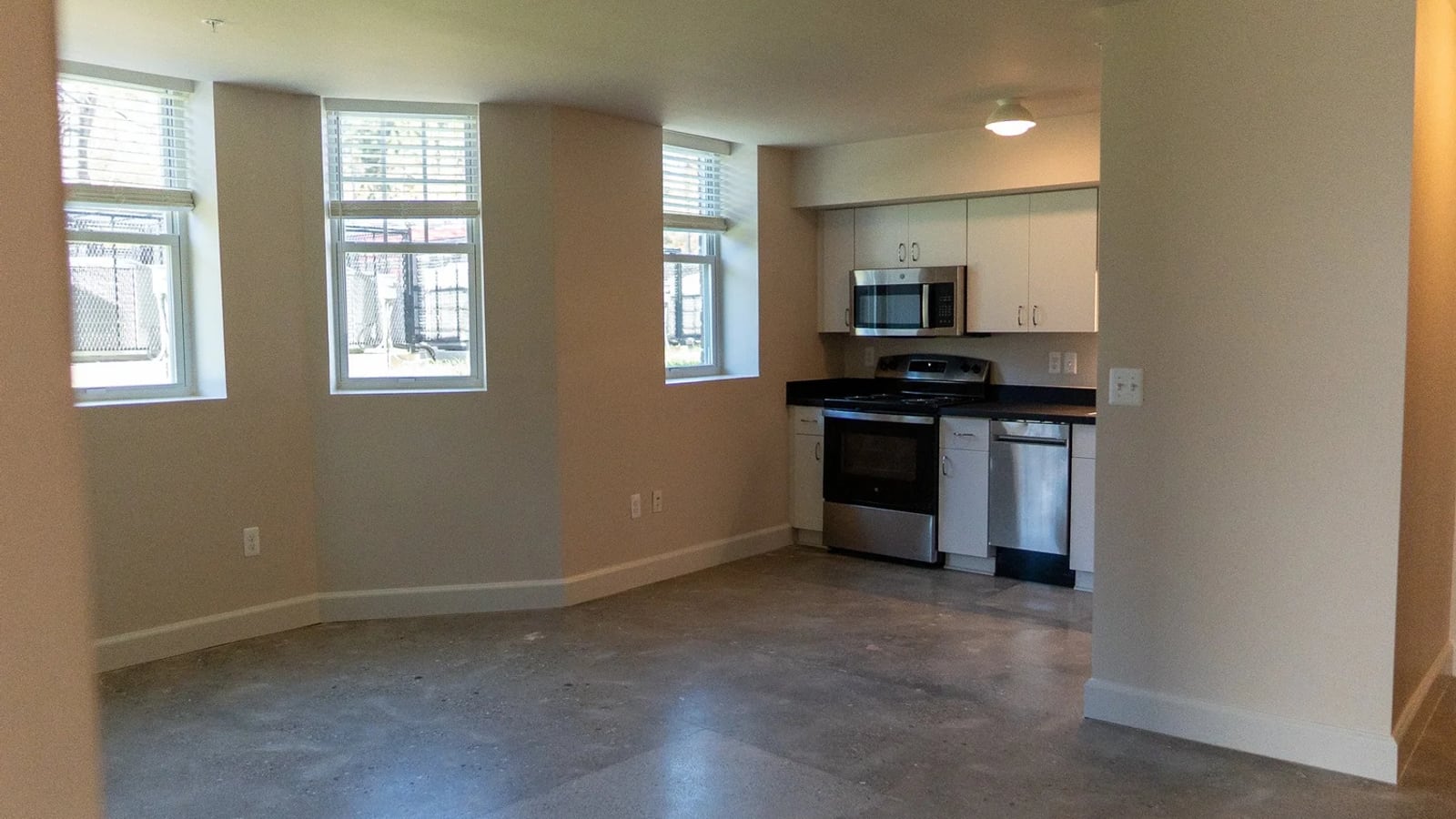 Photograph of an empty apartment with a sealed concrete floor, three windows and an open kitchen with a steel range and dishwasher