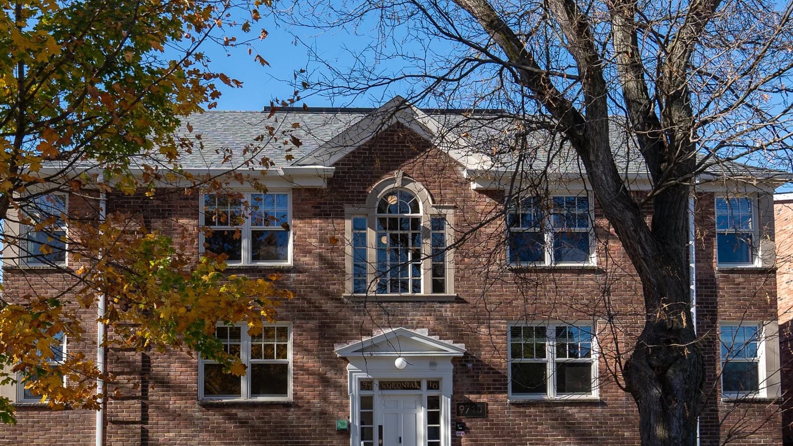 Photograph of the front of a three-story red brick apartment building with a white doorway, an arched window above it and bare autumn trees in front