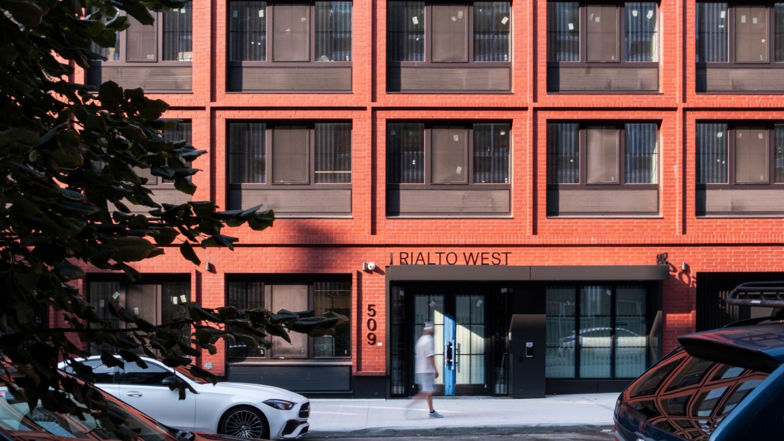 Photograph of the lower floors of a red brick building with dark-framed windows, metal lettering reading RIALTO WEST above a dark canopy over glass doors, the number 509 set vertically on the brick beside the entrance, a person walking past blurred by motion, parked cars in the foreground and leaves at the left edge