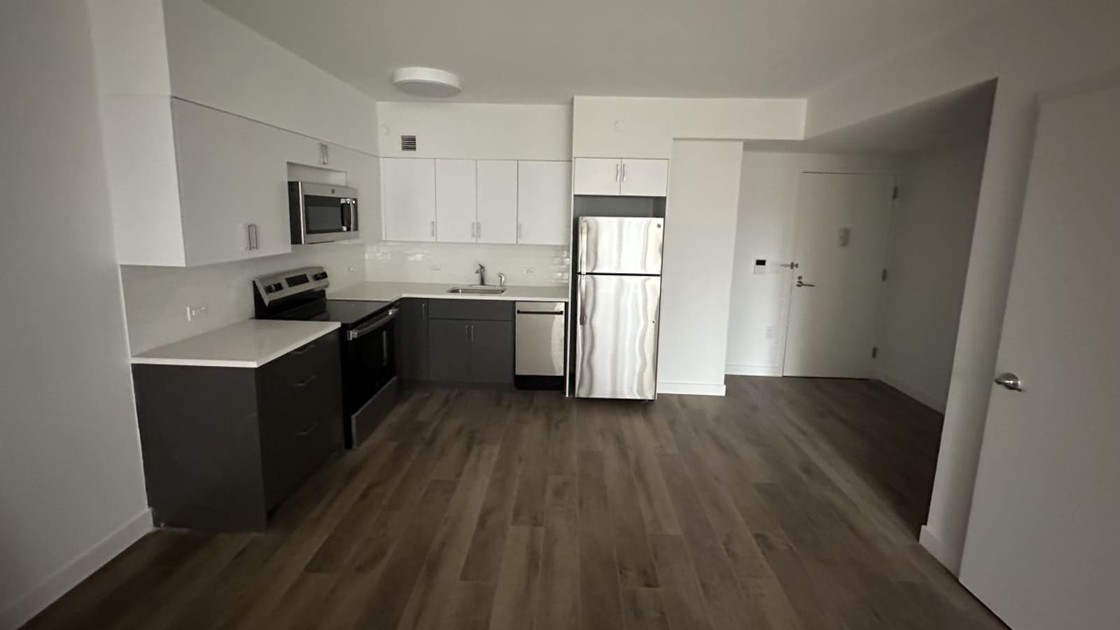 Photograph of an empty open kitchen with white upper cabinets, dark gray lower cabinets, a stainless steel range with a flat cooktop, a microwave, a dishwasher and a refrigerator, white tile backsplash and wood-look plank flooring, with the apartment entry door at right