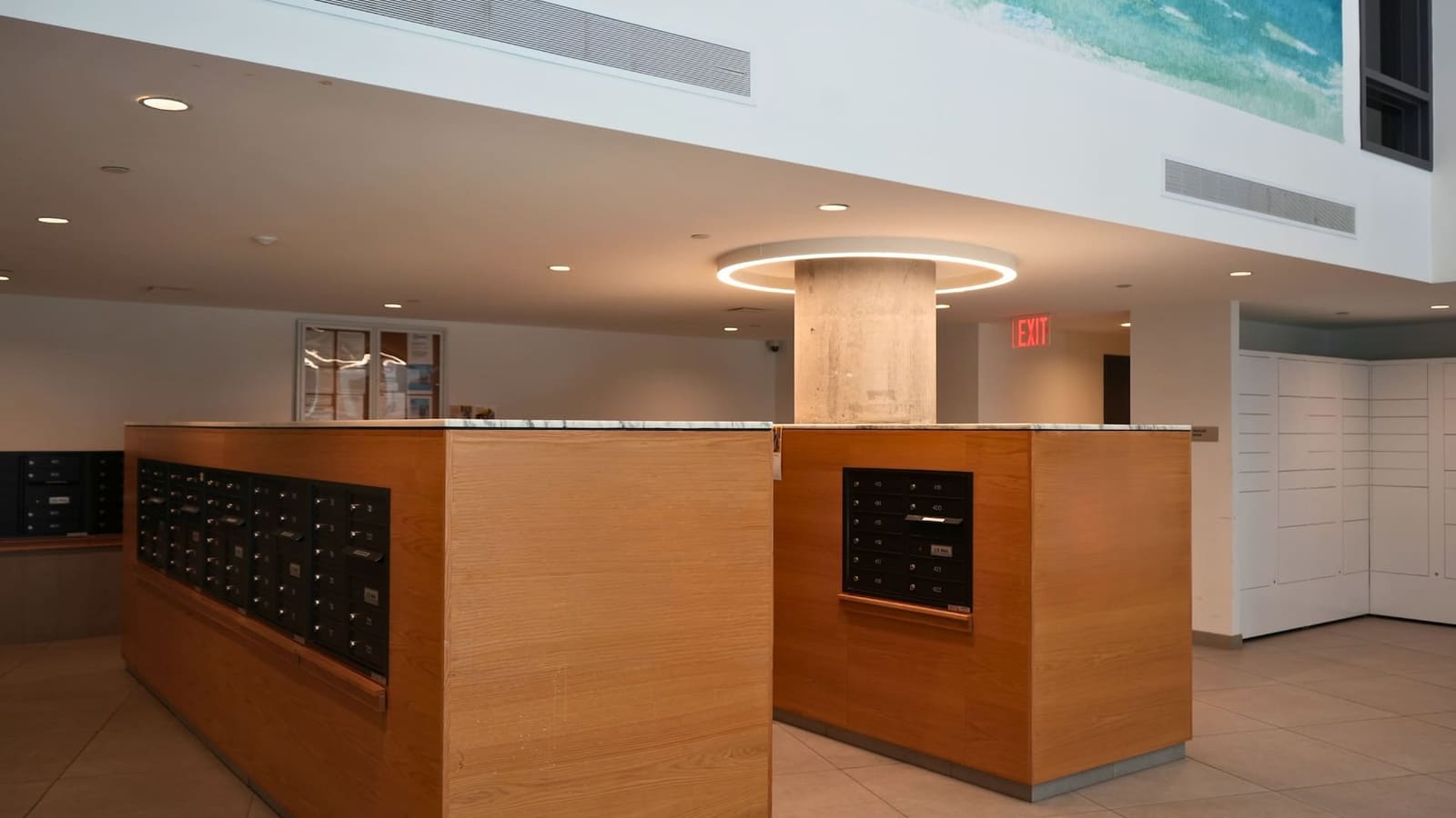 Photograph of a lobby with two wood-paneled mailbox islands topped with white stone, a round concrete column ringed by a circular light, white parcel lockers at right and a blue-green watercolor mural on the upper wall