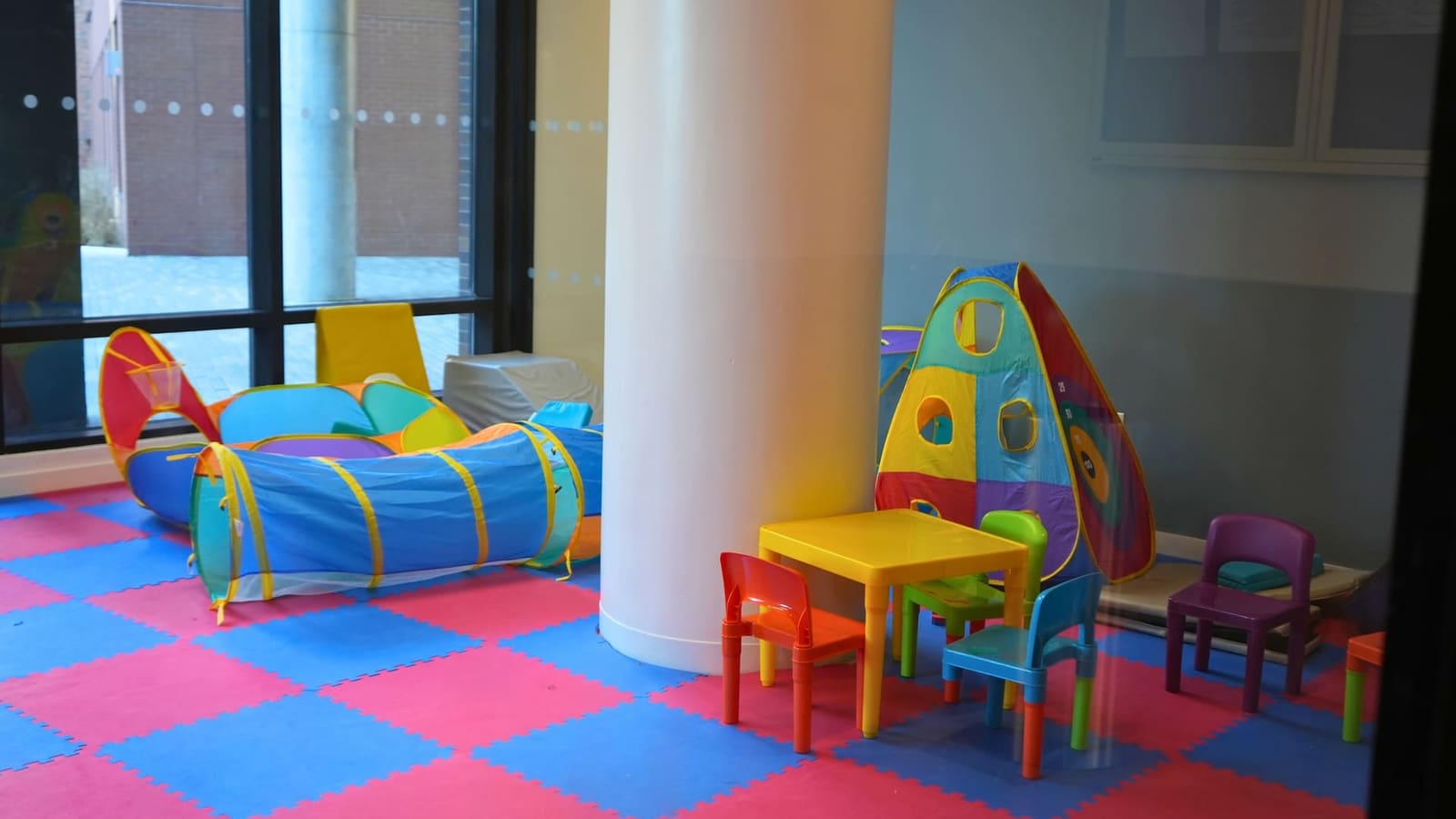 Photograph through a glass wall of a children's playroom with red and blue foam floor tiles, a play tunnel, a pop-up tent, a small yellow table with colored plastic chairs and a white round column