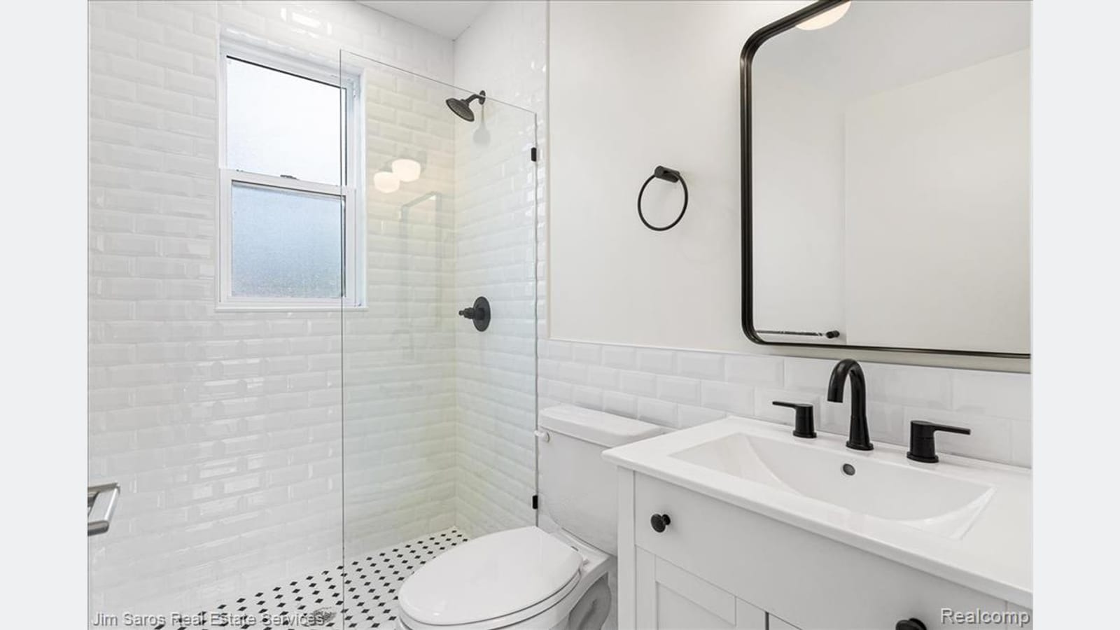 A bathroom with white tile walls, a shower behind a glass panel, a black and white patterned floor, a toilet and a white vanity with black fixtures