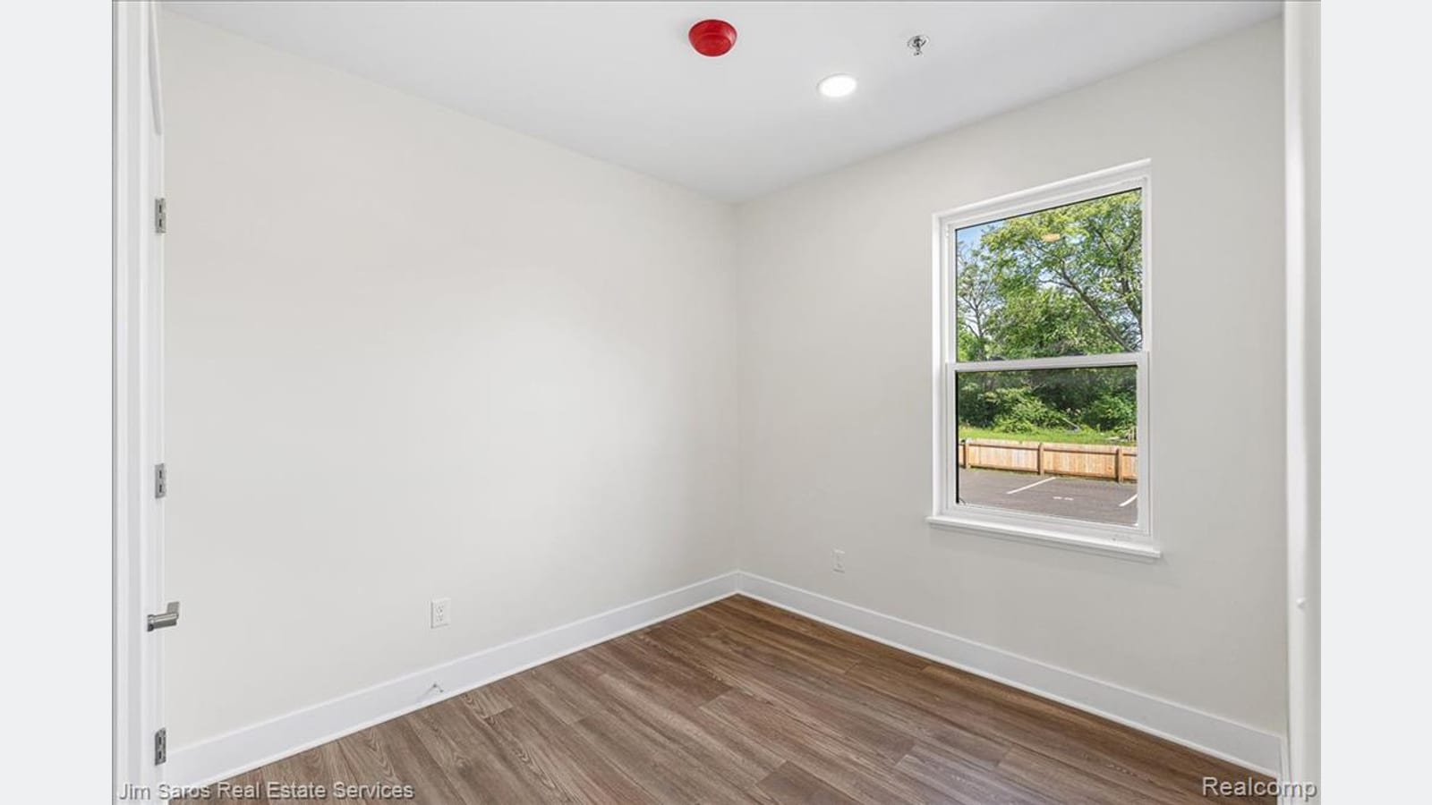 An empty bedroom with white walls, a wood-look floor and one window looking onto a parking area and trees