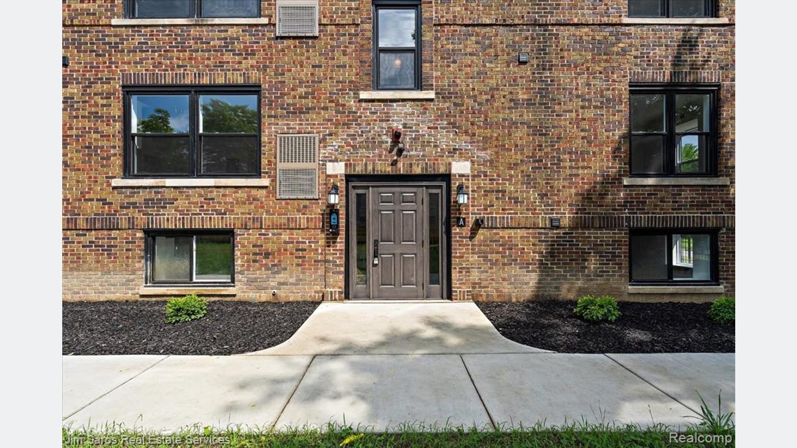 Photograph of a brick apartment building entrance with a dark door between two rows of windows, a concrete walk and new shrubs in mulch beds