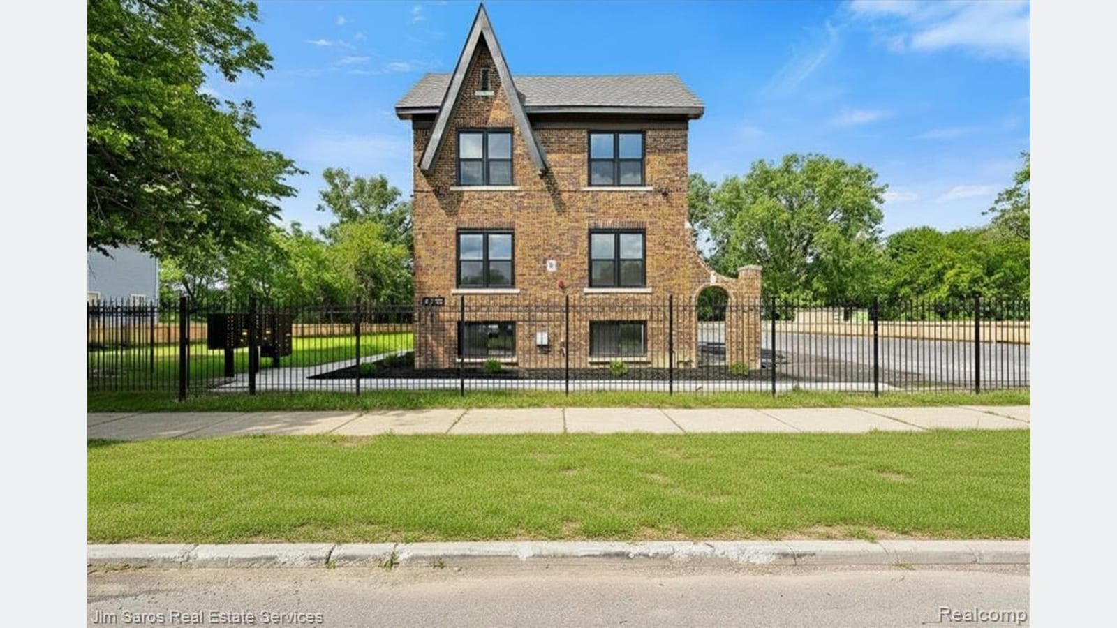 Photograph of the narrow gabled front of a brick apartment building with three rows of windows, behind a black metal fence and a lawn, with trees on both sides