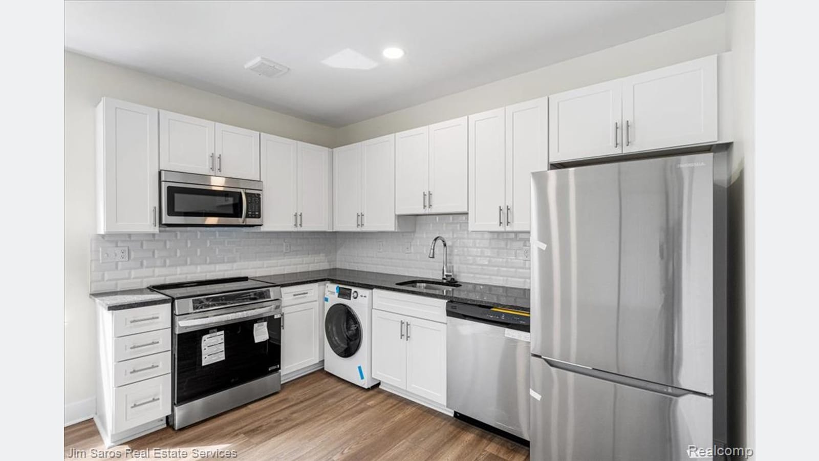 An apartment kitchen with white cabinets, dark stone counters, a white tile backsplash, a stainless steel refrigerator, range, microwave and dishwasher, and a front-loading laundry machine under the counter