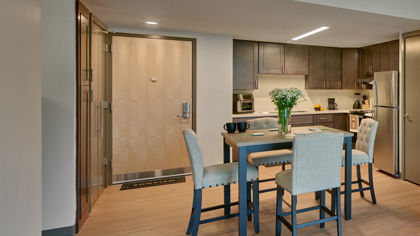 Apartment entry door beside a kitchen with dark wood cabinets, a stainless-steel refrigerator and range, and a dining table with four upholstered chairs on a wood-look floor