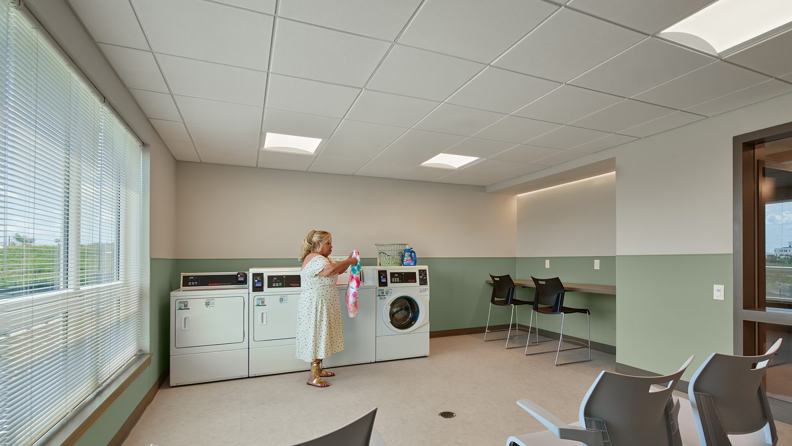Laundry room with white washers and dryers along a green-banded wall, a woman holding a garment, a counter with stools and a wall of windows with blinds