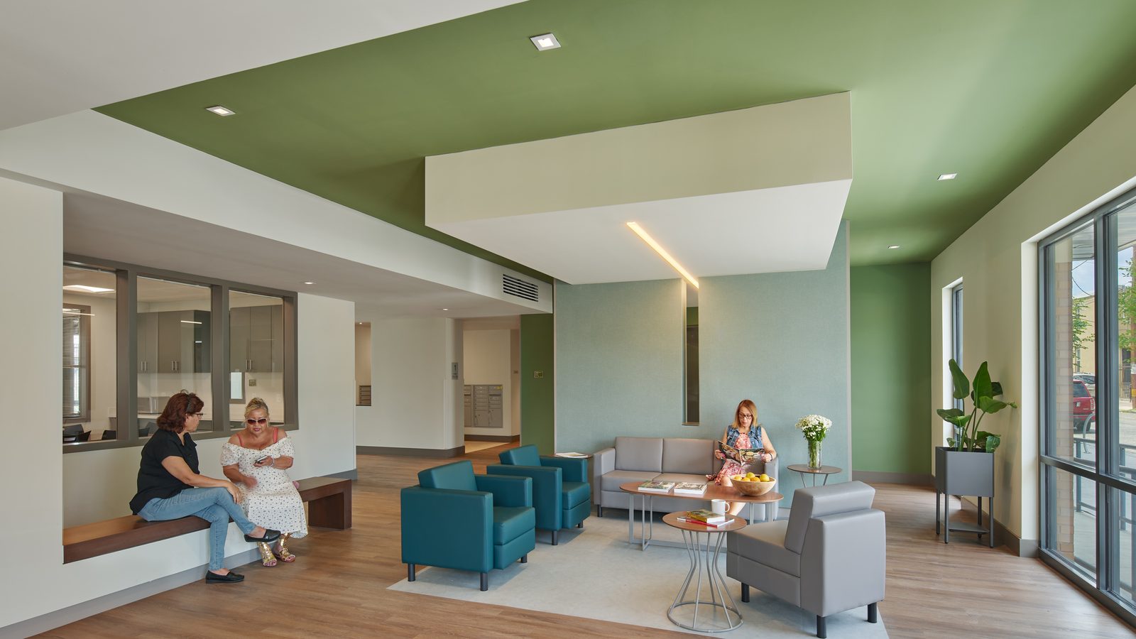 Lobby with a green ceiling panel, teal and gray armchairs around low tables, a bench along a window into an office, and three people seated