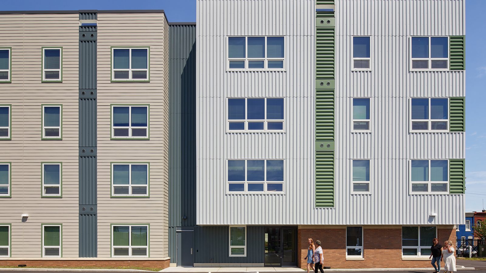 Rear of a four-story building in white ribbed metal, gray siding and brick with green louvered panels, a rear door and a striped parking lot where four people are walking
