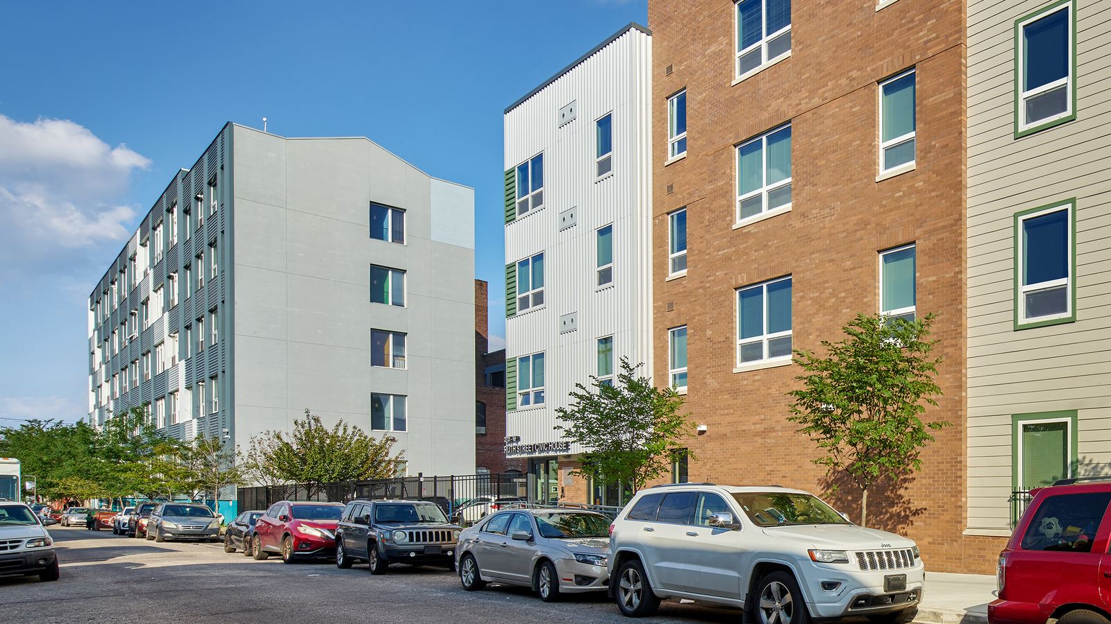 Street lined with parked cars beside a brick and white metal apartment building, with a taller gray and white building beyond it under a blue sky