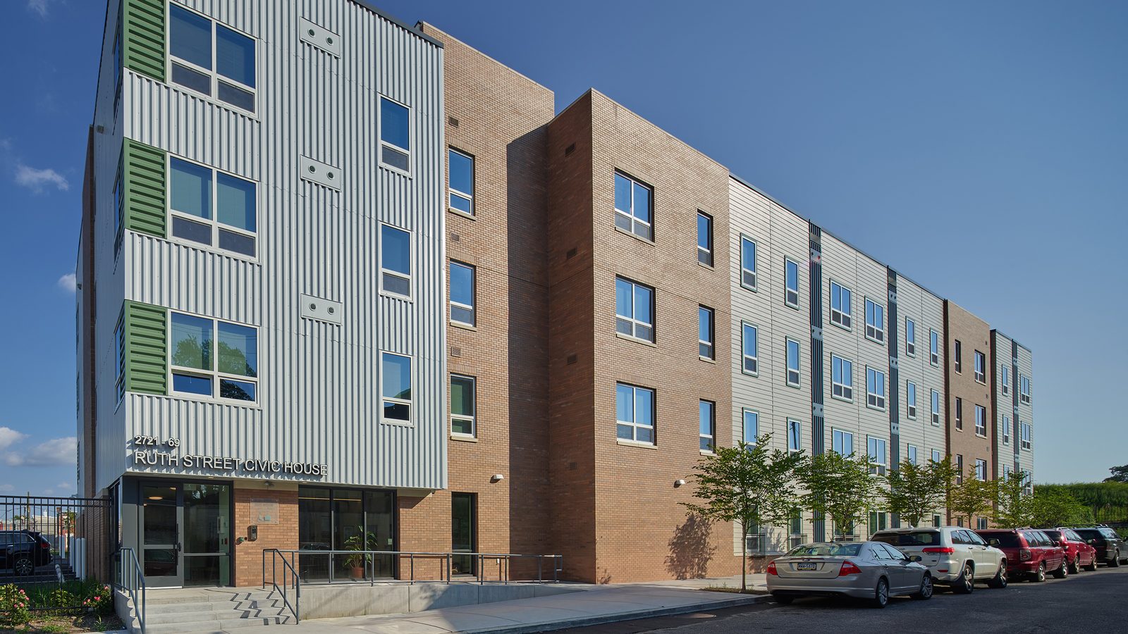 Four-story apartment building in white ribbed metal, green window panels and tan brick, with the name Ruth Street Civic House over a glass entrance and cars parked along the street