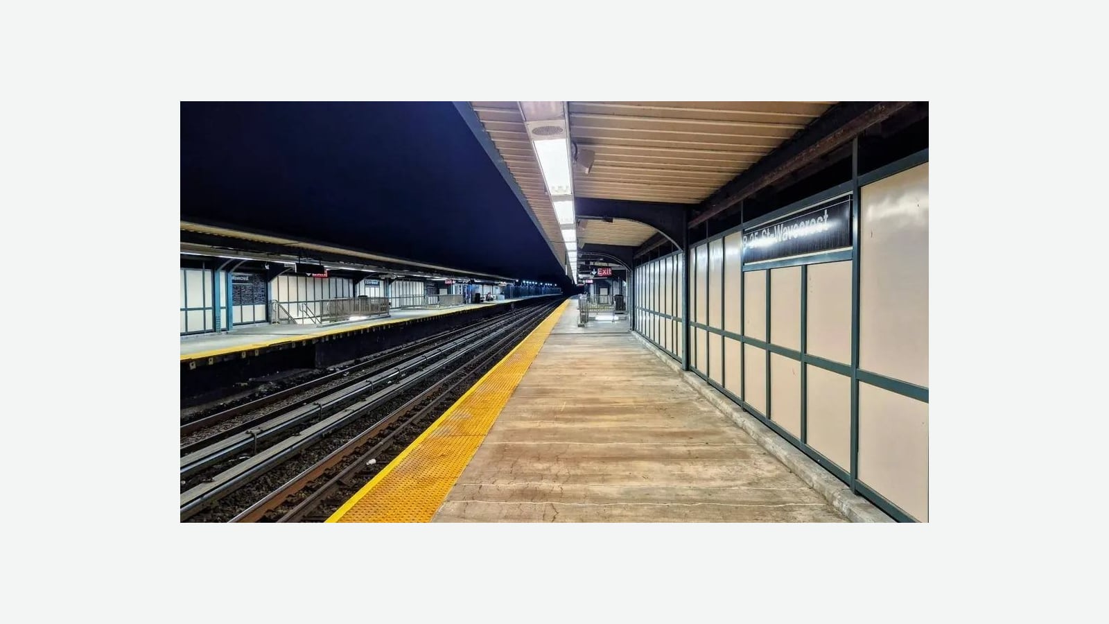 Photograph of an empty covered subway platform at night with a yellow edge strip, tan wall panels in green frames, a sign reading B 25 St-Wavecrest at right, a red Exit sign in the distance and a second lit platform across the tracks at left, shown at its original size on a light gray background