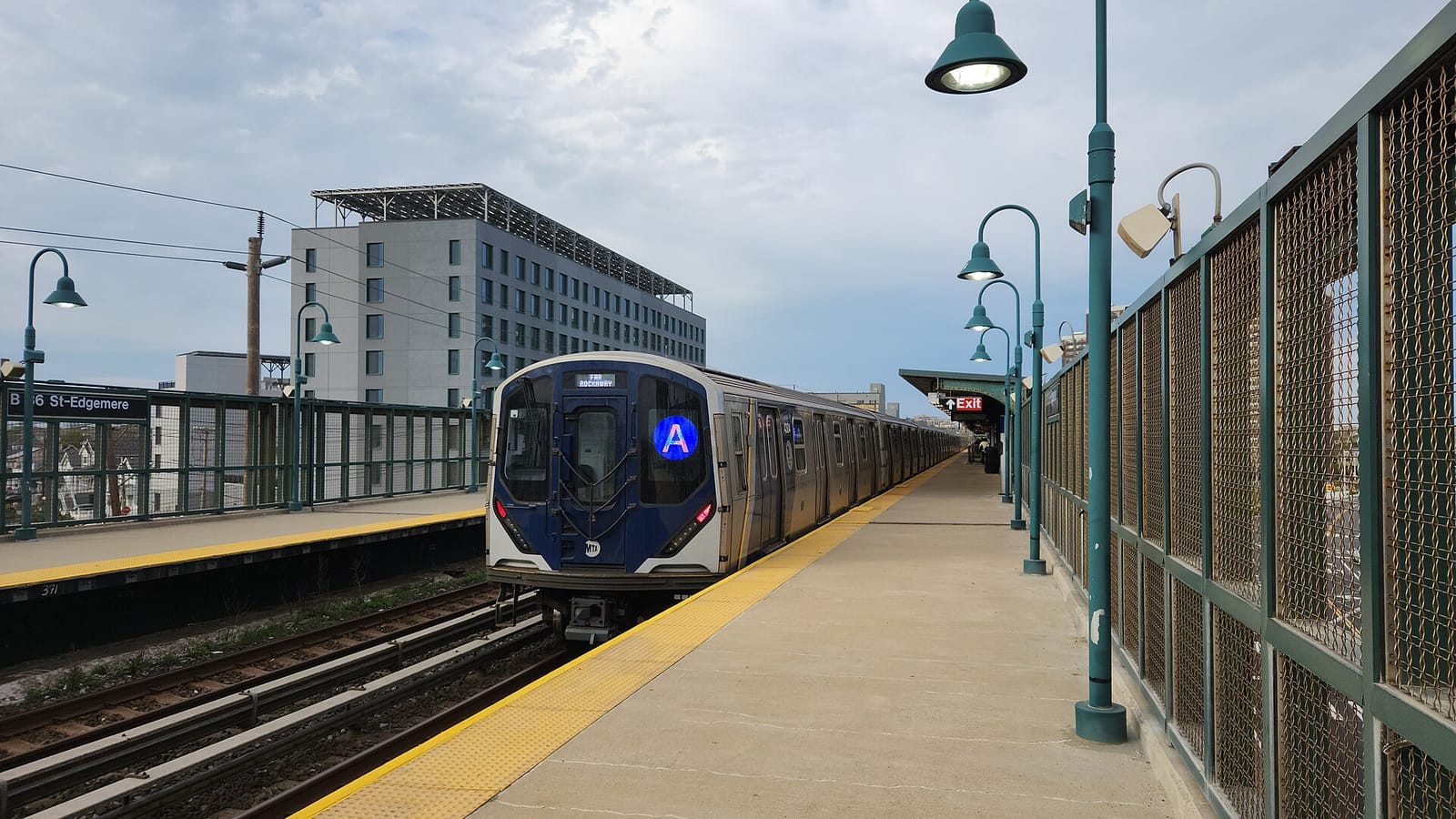Photograph along an elevated open-air subway platform with a yellow edge strip and green lamp posts, a blue and silver train with a lit A sign stopped at the platform, a station sign reading B 36 St-Edgemere at left, and a gray multistory building with solar panels on its roof behind, under a cloudy sky