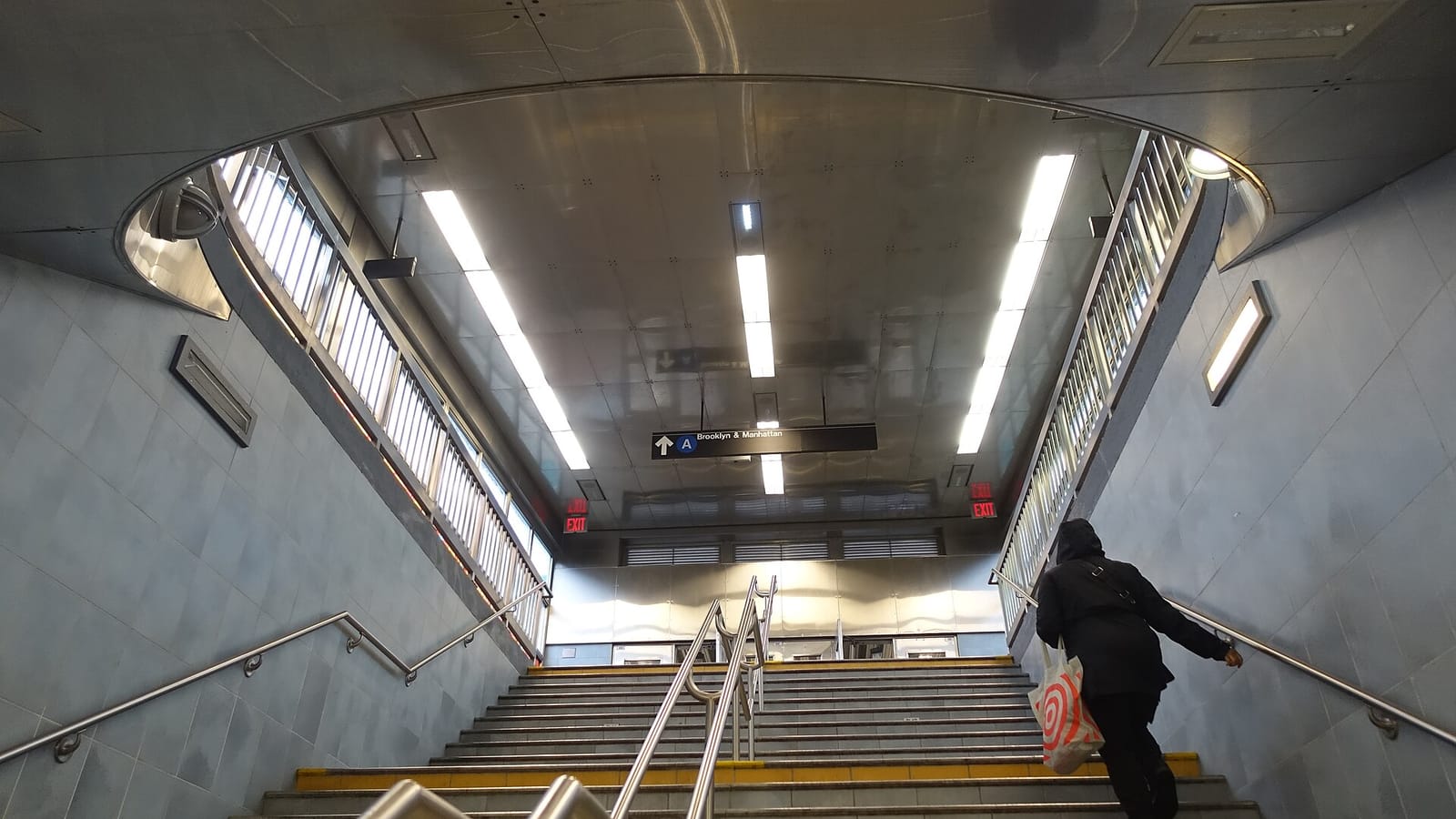Photograph up a wide indoor stairway with metal handrails, gray tiled walls and a curved stainless-steel ceiling with long lights, a sign reading A Brooklyn and Manhattan with an up arrow, red exit signs, and one person seen from behind climbing the stairs with a bag