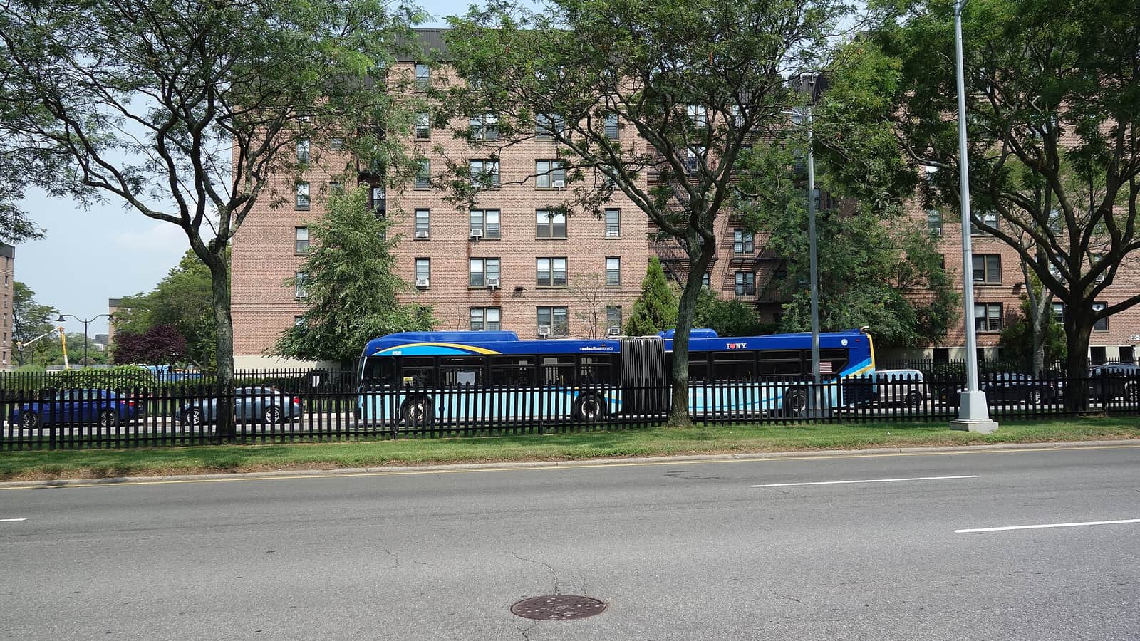 Photograph across a wide, empty street with a grass strip and a black picket fence, a long blue articulated bus parked or moving behind the fence, and a seven-story red-brick apartment building with window air conditioners behind leafy trees, under a hazy sky