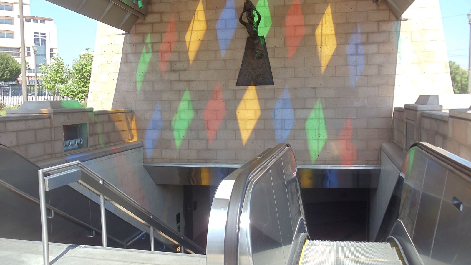 Escalators and stairs leading down into the Mariachi Plaza rail station, with coloured light from the canopy on a stone wall