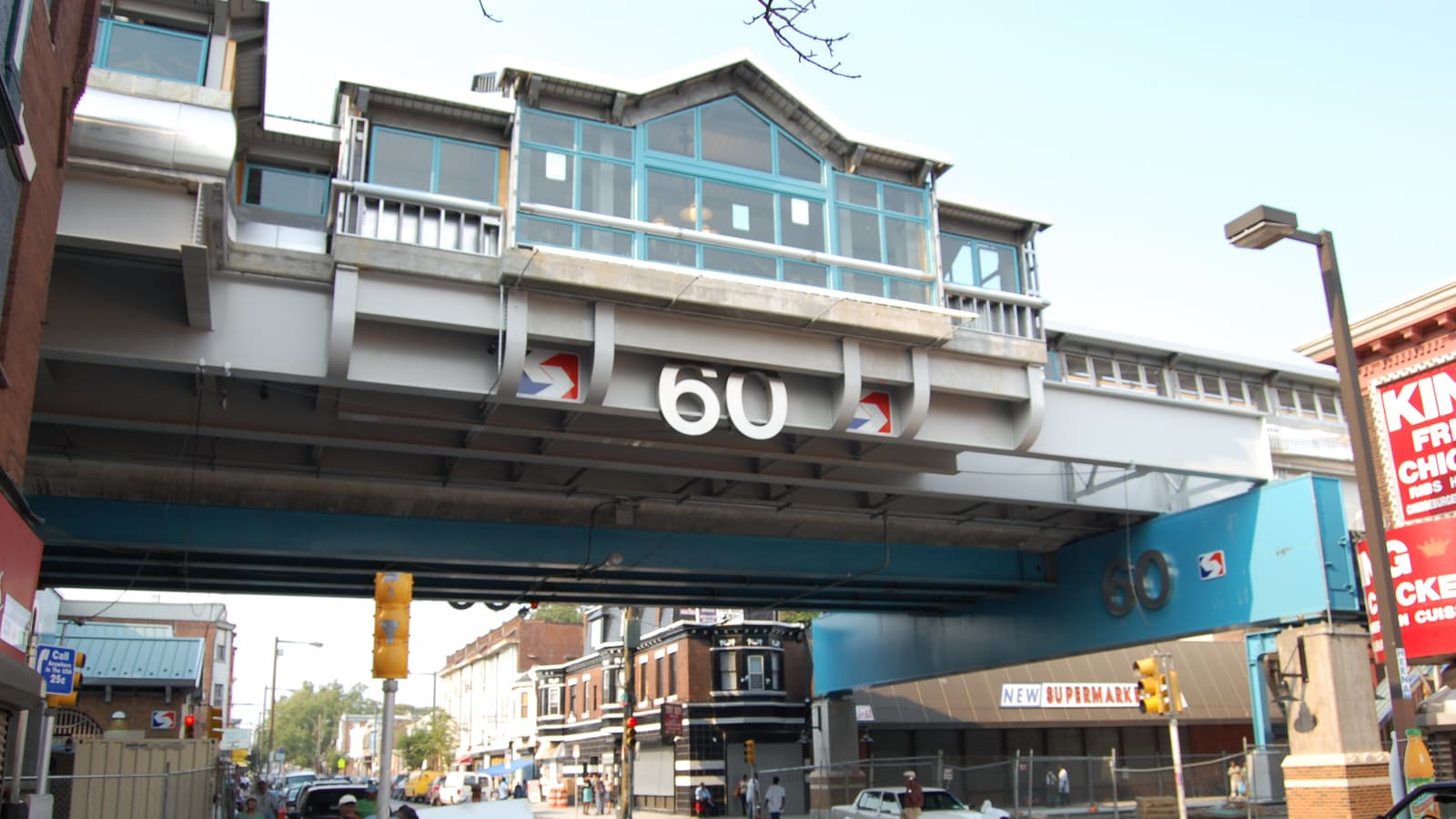 An elevated railway station of glass and grey steel spanning a city street, with a large number 60 on the girder