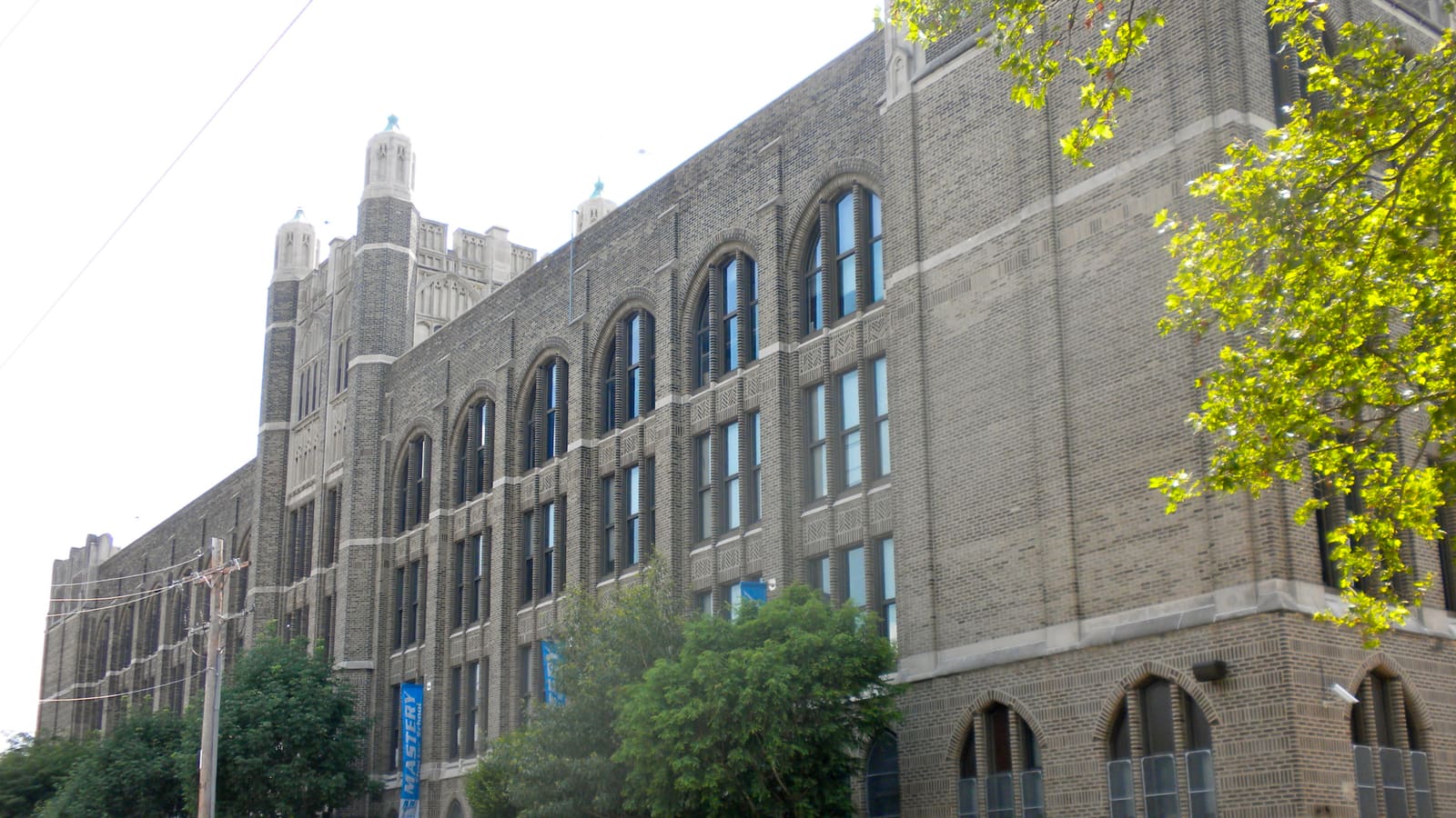 A long four-storey school of grey-brown brick with tall arched windows and a turreted stone entrance tower, seen from the street