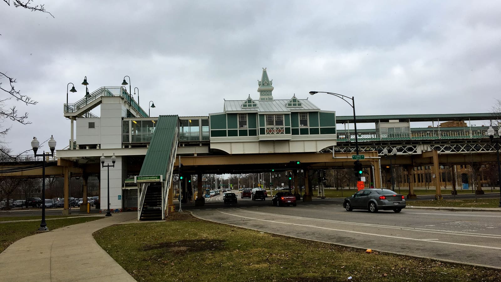 An elevated train station with a green and white Victorian-style station house over a street, with stairs signed Conservatory and cars passing beneath