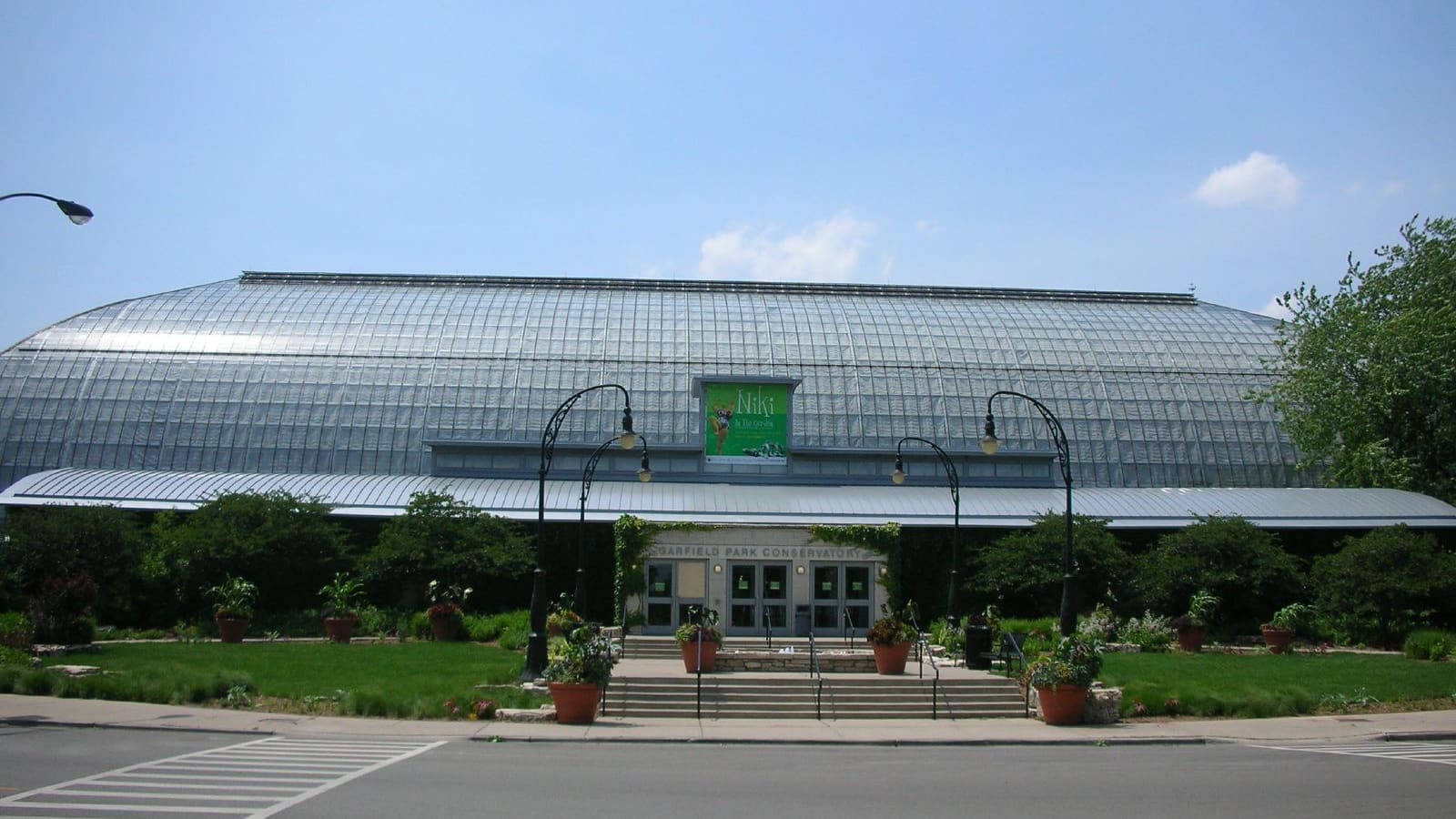 A long, low glass-roofed building with a sign reading Garfield Park Conservatory over its entrance doors, behind lawn, steps and planters