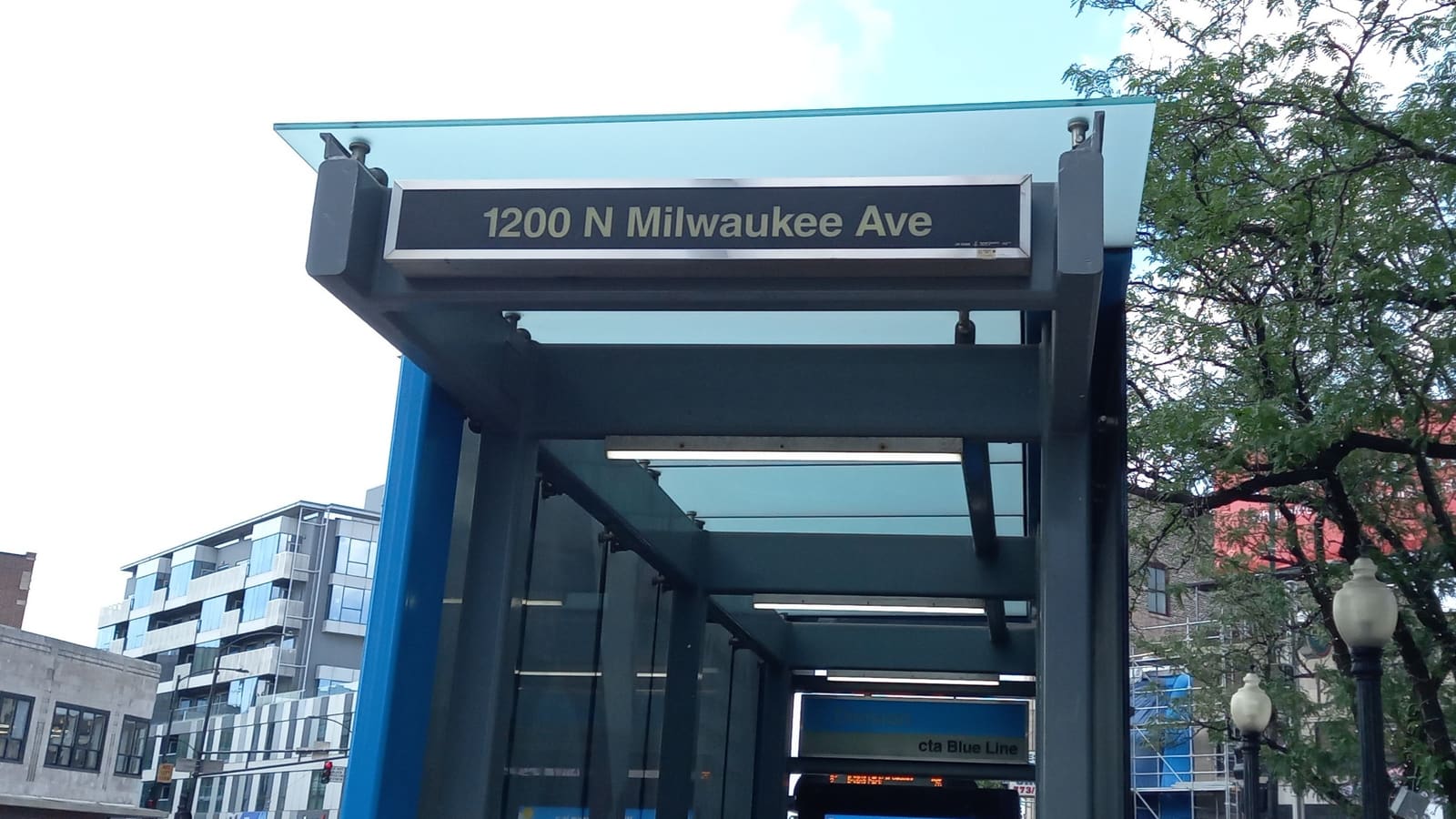 A glass and steel canopy over a subway entrance with a sign reading 1200 N Milwaukee Ave and a CTA Blue Line sign below it
