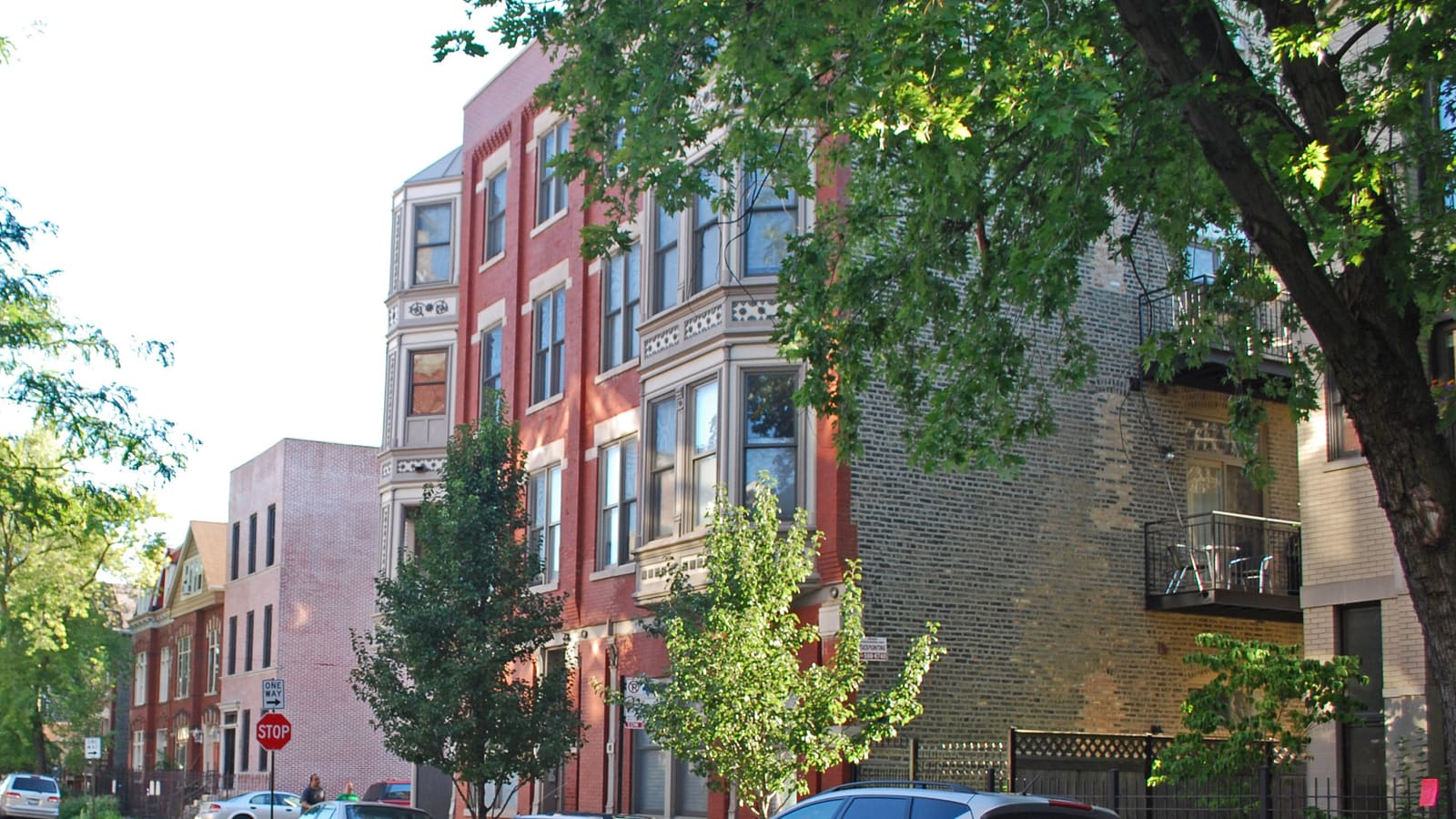 A four-storey red brick flat building with stone trim and bay windows on a tree-lined street, with older brick houses beyond it