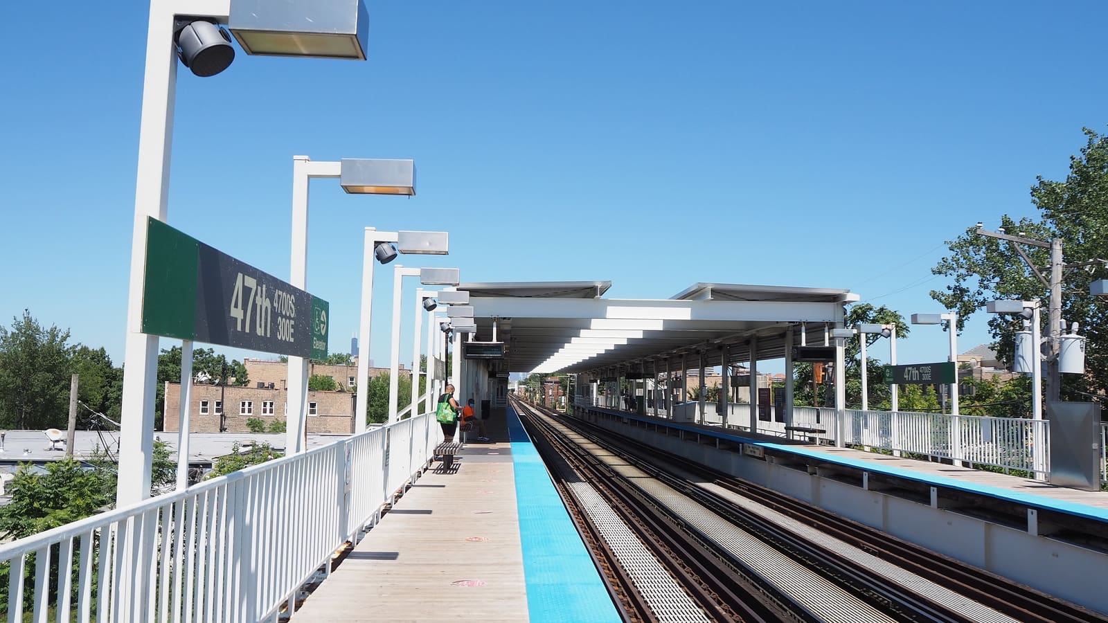 An elevated train platform with white canopies and a green station sign reading 47th, with brick buildings and trees beyond