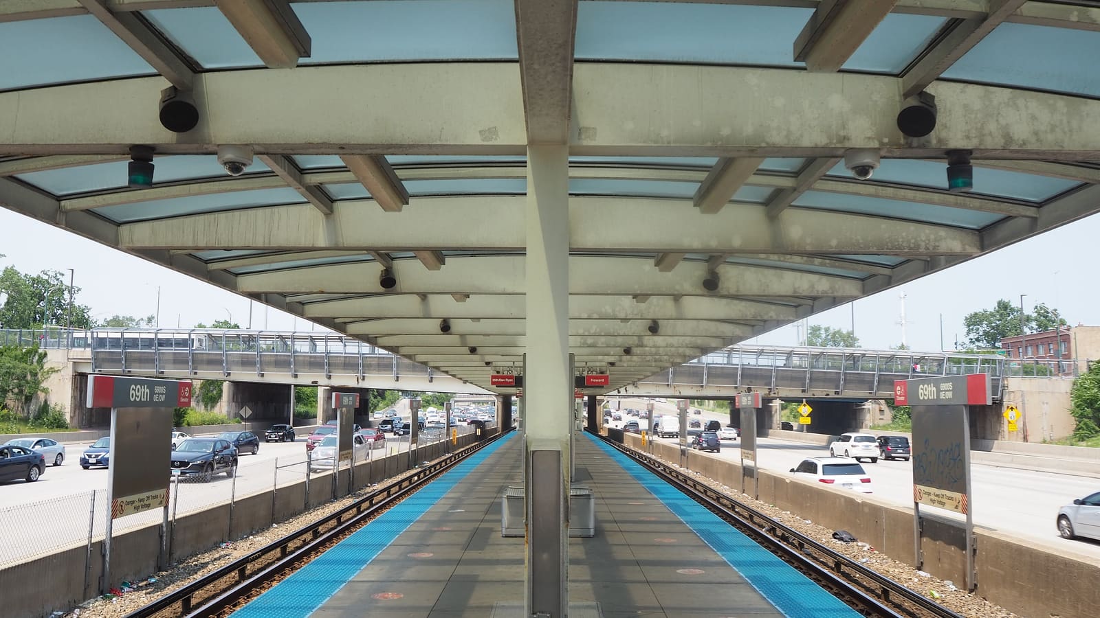 A train platform under a concrete and glass canopy in an expressway median, with red signs reading 69th and traffic on both sides