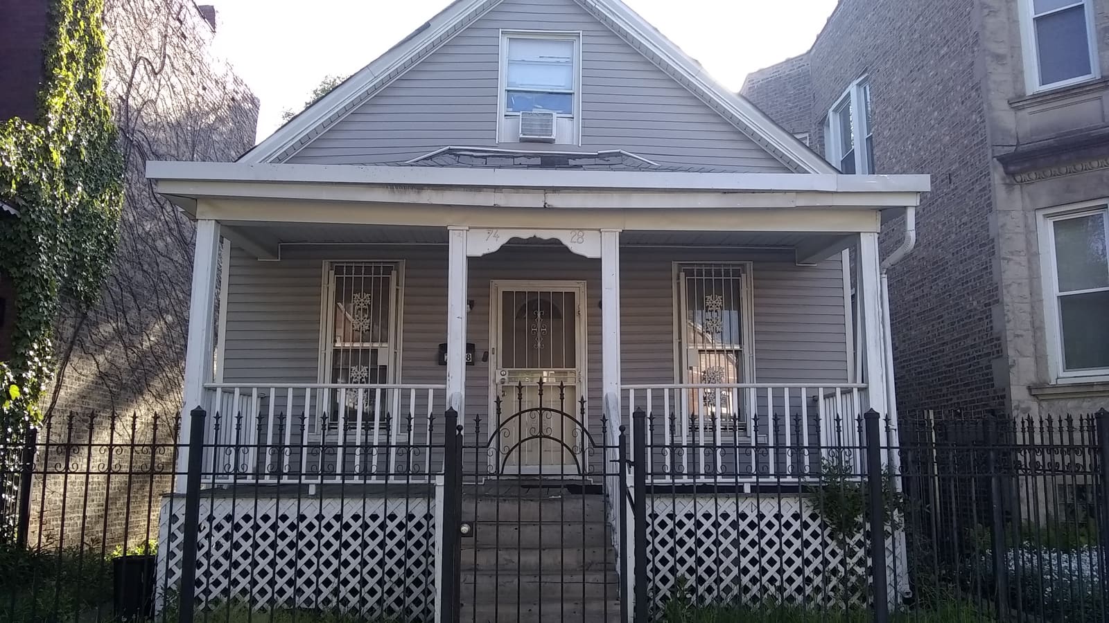 A grey frame house with a front porch and gabled roof behind an iron fence, between two brick buildings