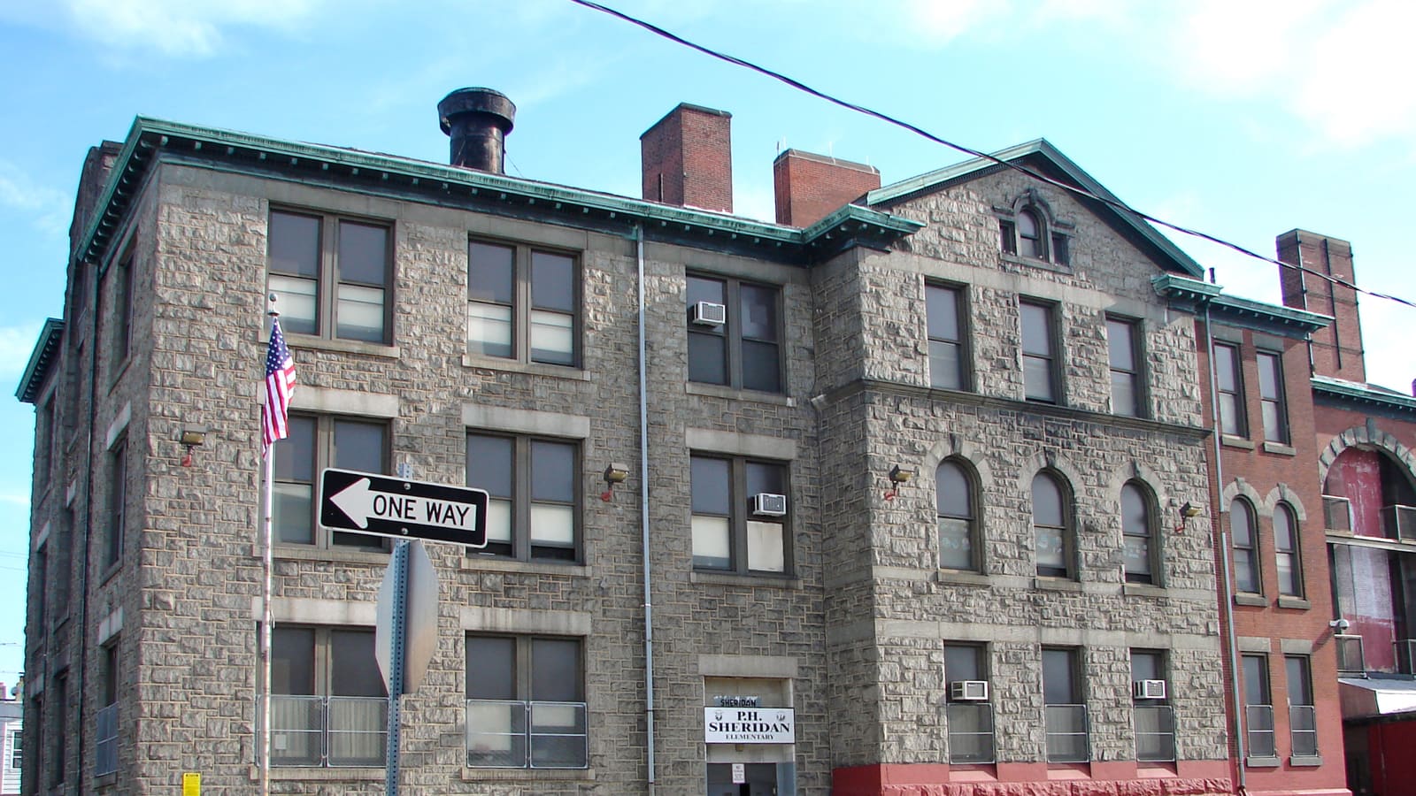 A three-storey school of rough grey stone with a green copper cornice and brick chimneys, and a one-way sign in front