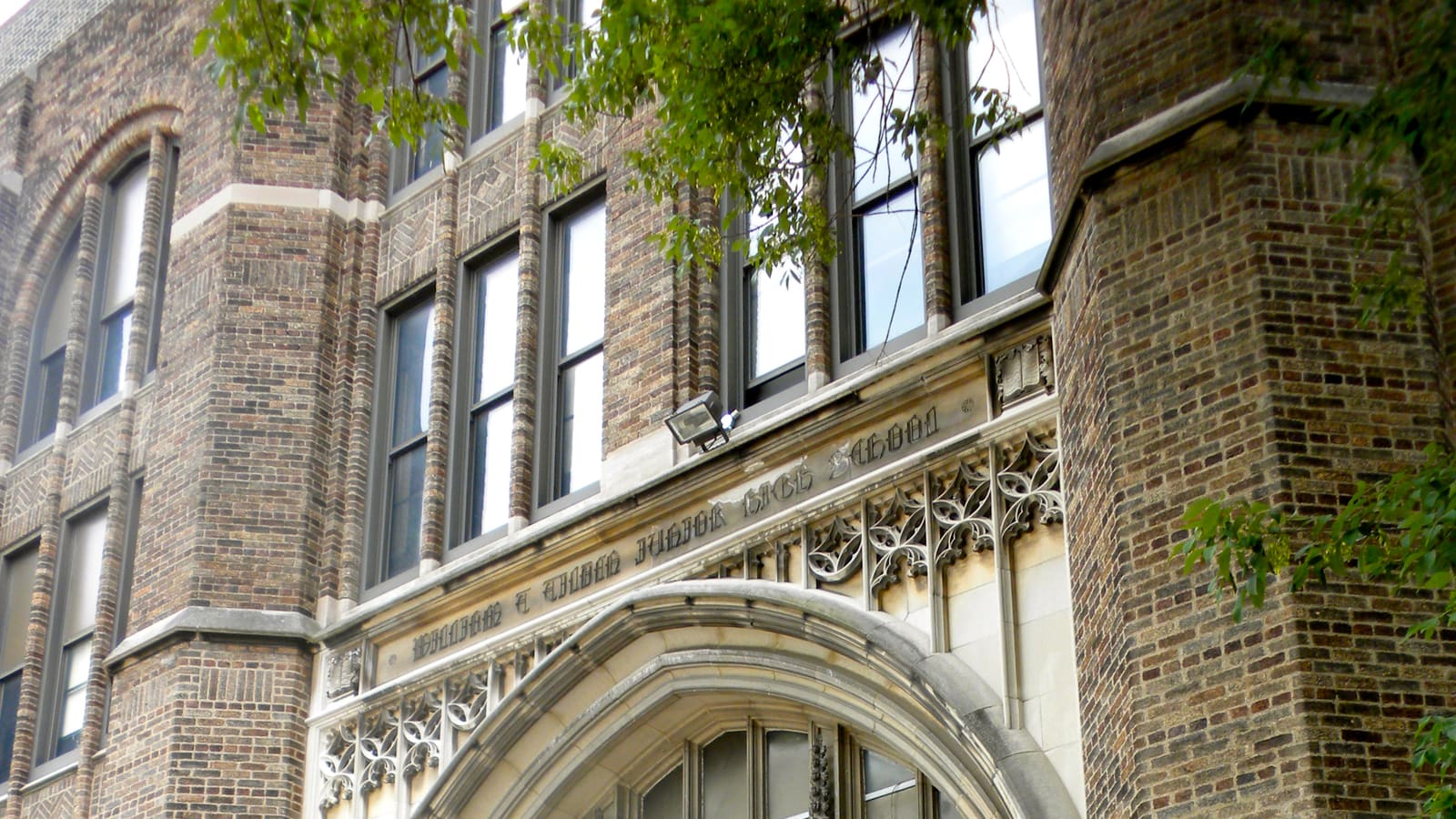 The brick and carved-stone front of a school, with tall windows above a Gothic arch and the school's name cut into the stone