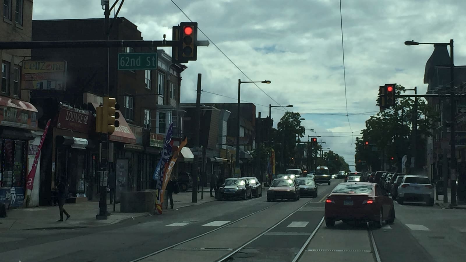 A shopping street with trolley rails set in the roadway, overhead wires, a 62nd St sign on the signal arm and shops along the left side