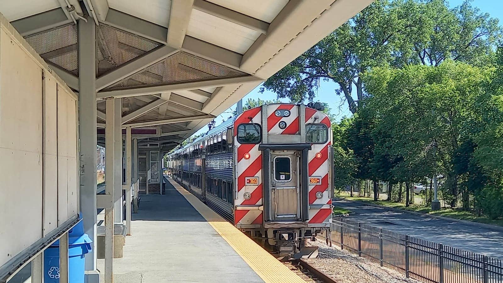 A double-deck commuter train with red and white striped end standing at a covered platform beside a tree-lined street