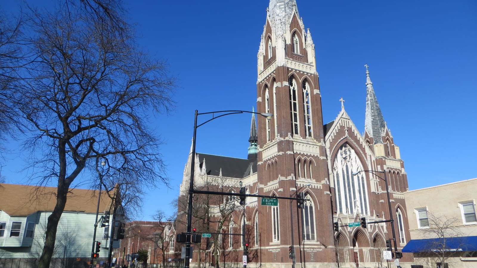 A tall red brick Gothic church with a central tower and spires at a street corner under a clear blue sky