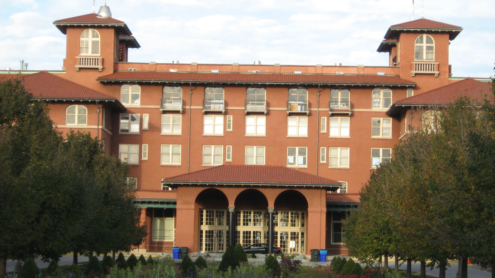 A large terracotta-coloured building with red tile roofs, two towers and an arcaded entrance behind trees and flower beds
