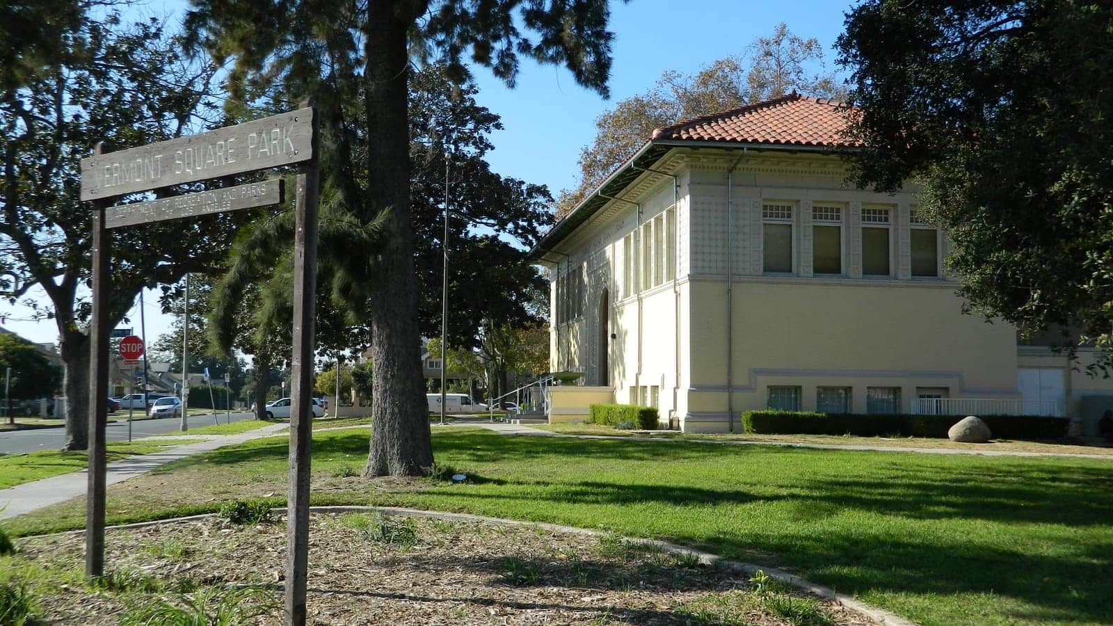 A cream brick library building with a red tile roof behind a wooden Vermont Square Park sign and a lawn