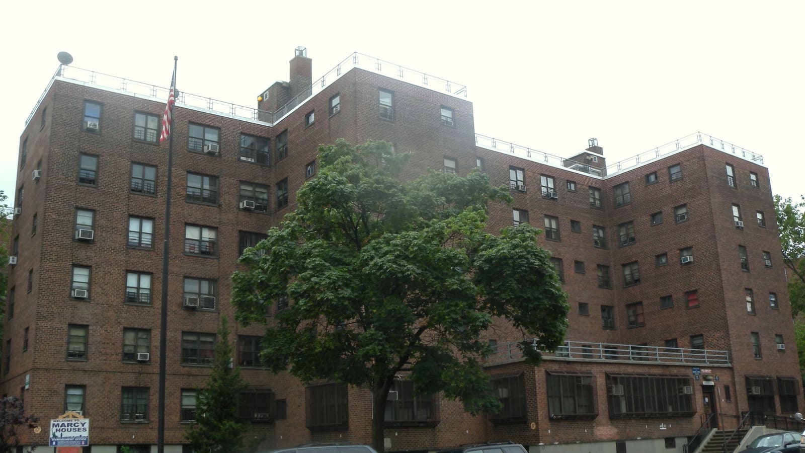 Six-story red-brick public housing buildings behind a large green tree, with a flagpole and a sign reading Welcome to Marcy Houses.
