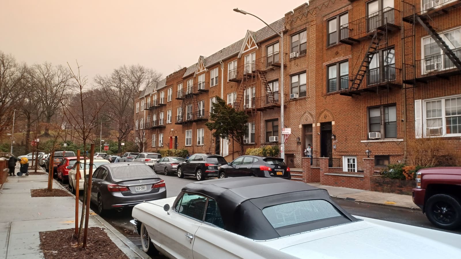 A quiet residential street lined with three-story red-brick apartment buildings with fire escapes, parked cars along both curbs and bare winter trees.