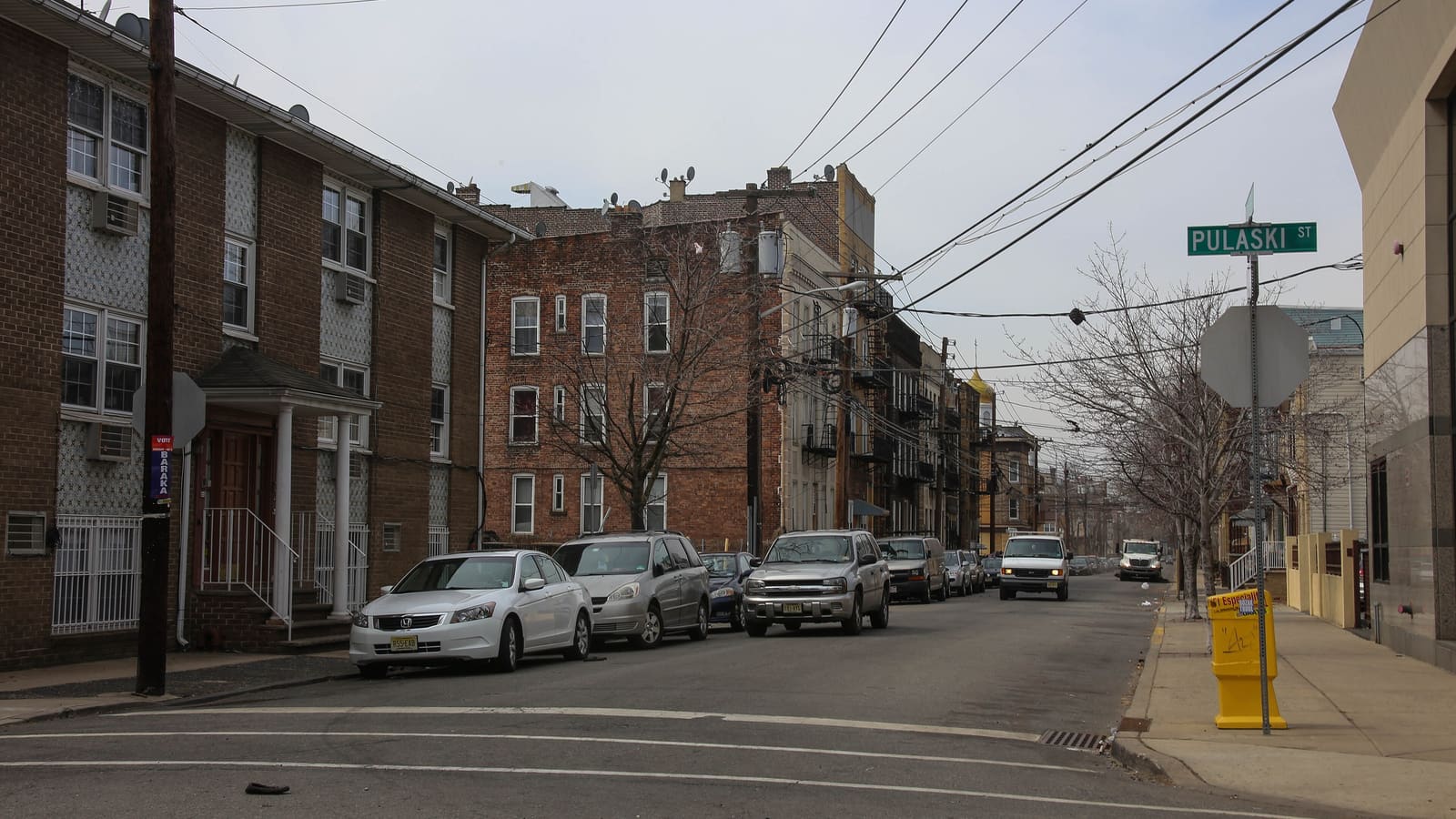 A street corner with a three-story brick garden apartment building on the left, older brick walk-up buildings beyond it, parked cars and a green Pulaski St sign