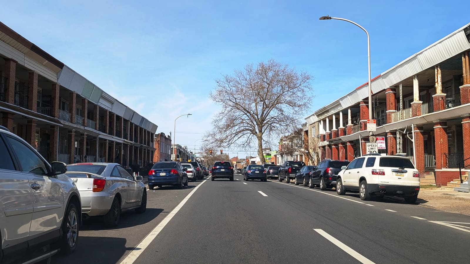A wide city street lined on both sides with two-story brick rowhouses with covered porches, parked cars along each curb and a bare tree at the end of the block