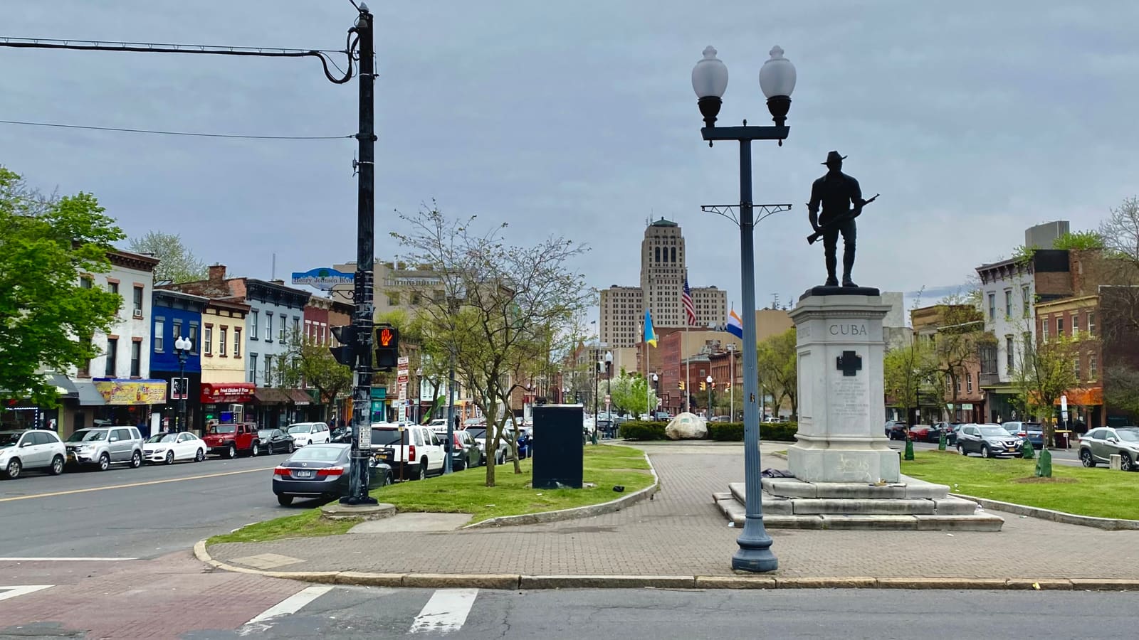 A small triangular park with a soldier statue on a stone pedestal and a twin-globe lamppost, rowhouses with shops on the left and an office tower in the distance