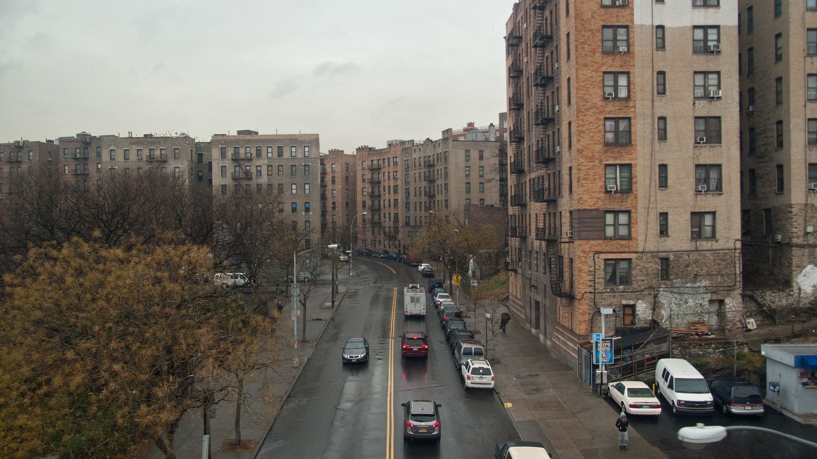 A wet two-way street curving between rows of six-storey tan and grey brick apartment buildings with fire escapes, cars parked along the curb and bare trees on one side