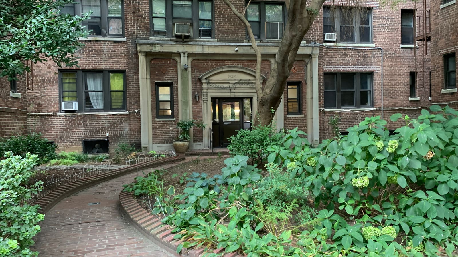 The entrance of a red-brick apartment building with a stone door surround, reached by a curved brick path through a planted courtyard with a tree and hydrangeas