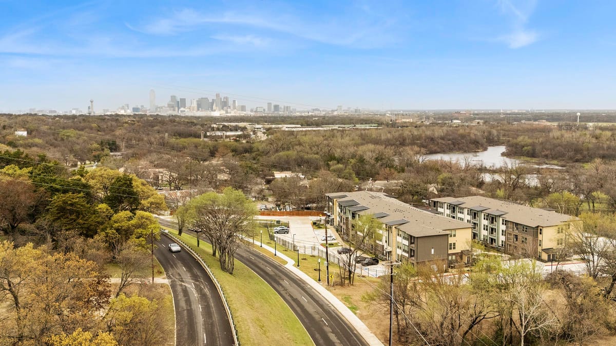Aerial view of two apartment buildings beside a curving divided road, surrounded by trees, with a pond behind them and the downtown Dallas skyline on the horizon