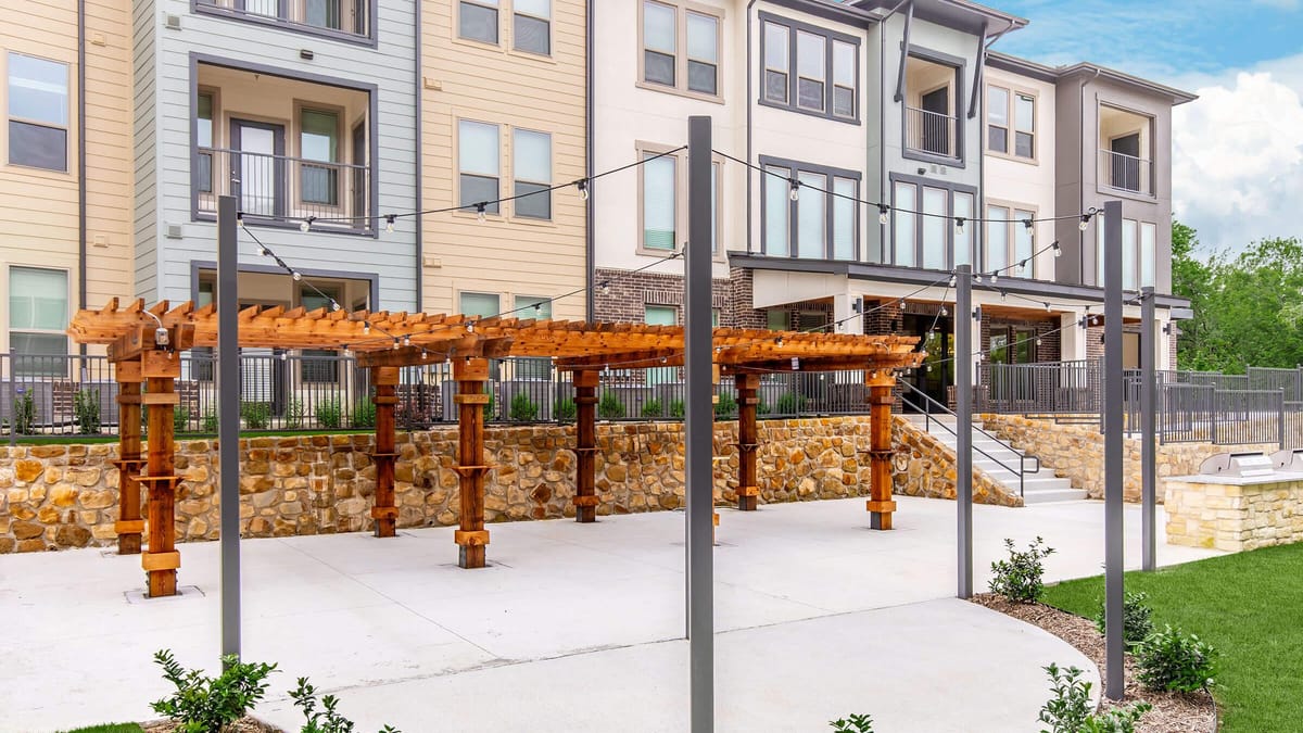 Wooden pergolas with string lights over a paved terrace and stone retaining wall in front of a three-story apartment building