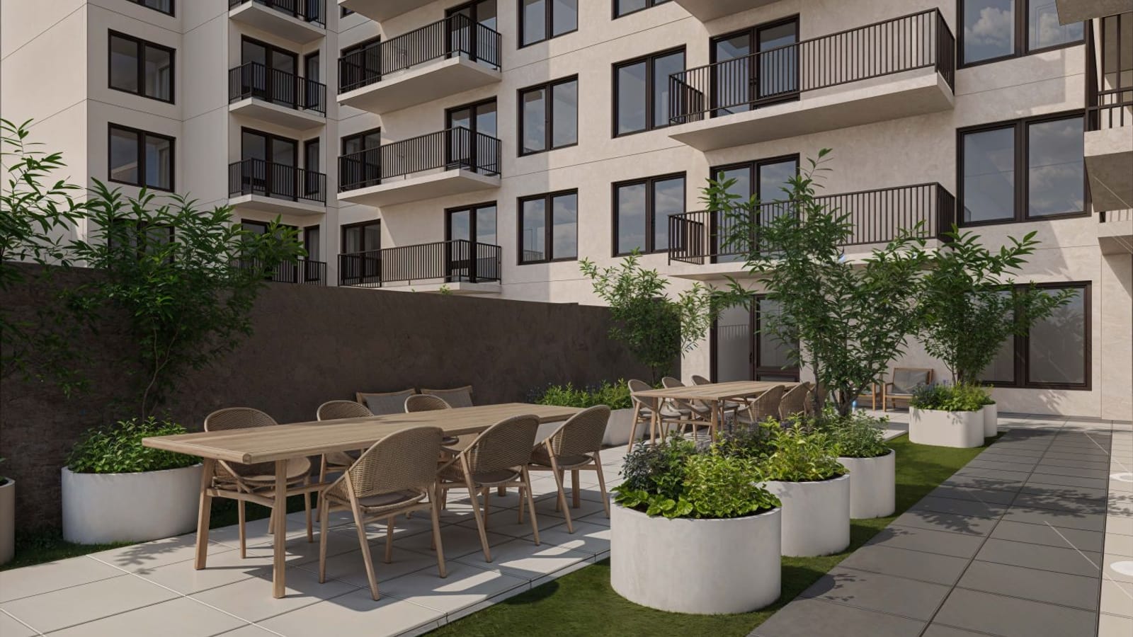 Rendering of a paved outdoor area with two long wood dining tables and woven chairs, round white planters with shrubs and small trees, a strip of lawn and a path, beside a pale building wall with dark-framed windows and balconies with dark railings